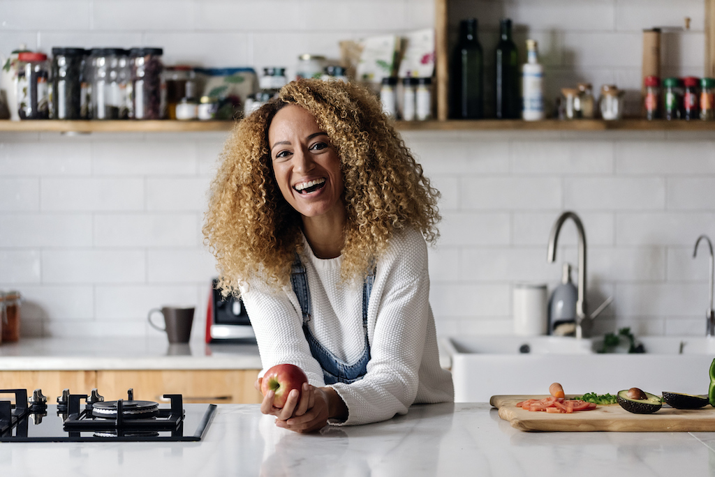 Stock photo of a middle aged woman holding an apple in a kitchen. She is smiling and looking at camera draining cleaning