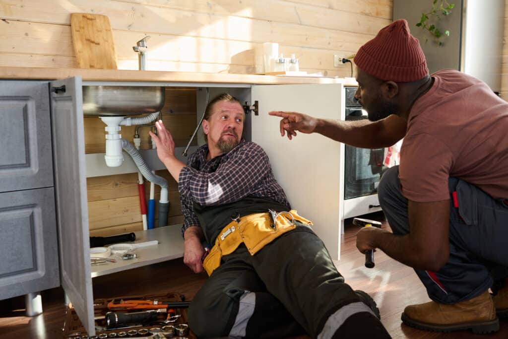 Common Plumbing Emergencies, Mature plumber looking at young colleague pointing at pipe under metallic sink in kitchen cabinet while explaining the problem.